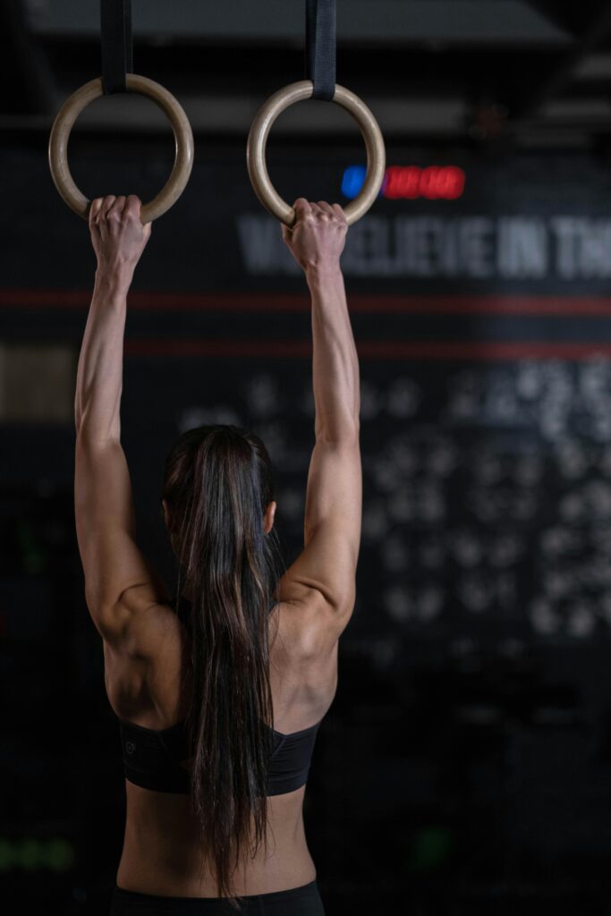 pexels-photo-7674484-7674484 Back view of a woman exercising on gymnastic rings in a dimly lit gym setting, showcasing strength.